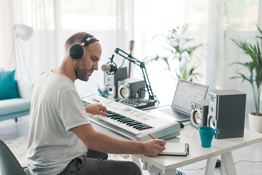 A focused musician wearing headphones, sitting at a keyboard in a bright, minimalist home studio. He is writing notes in a notebook while working on a music production setup, with a microphone, speakers, and a laptop in front of him.