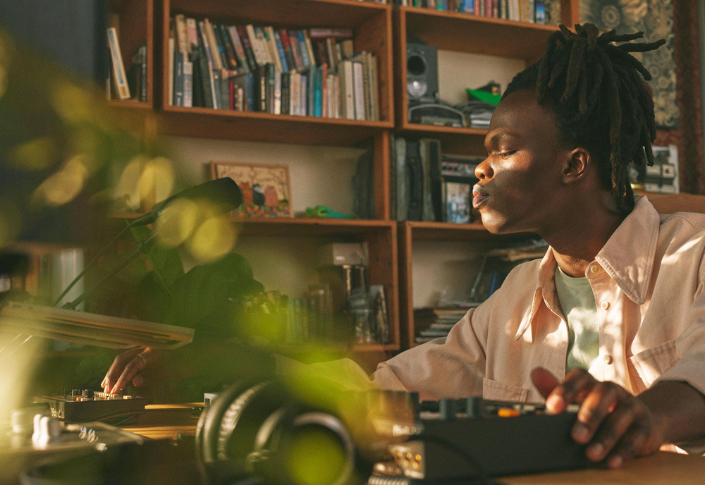 A musician with dreadlocks sits in a cozy, sunlit room filled with bookshelves, working on a music production setup with headphones and mixing equipment.